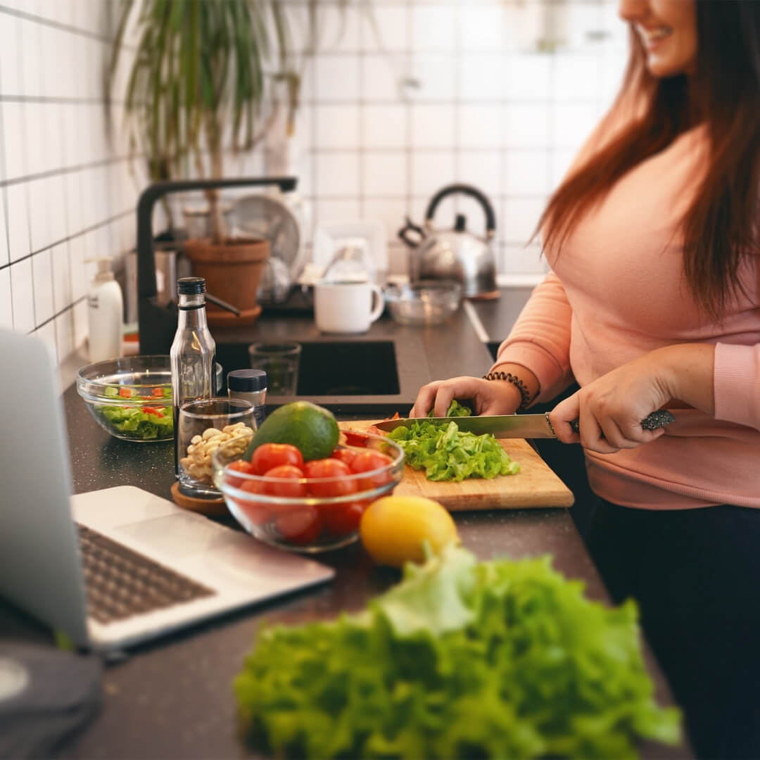A woman chopping vegetables in her kitchen as she looks at her laptop. Image is for the blog post about How to Get Started with the 4 Pillars of Weight Loss