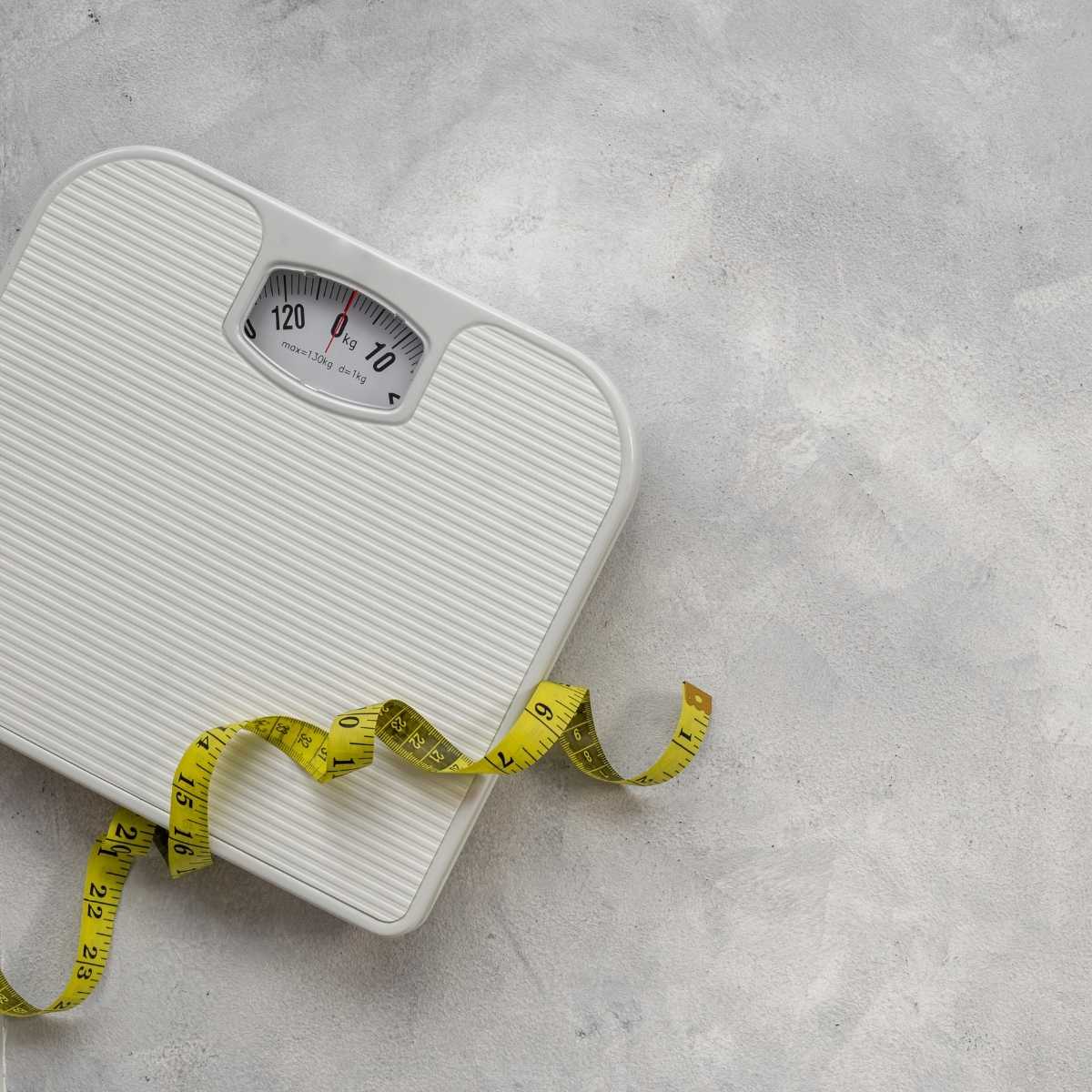 Image of a white bathroom scale with a yellow measuring tape draped across it, placed on a light gray textured surface.