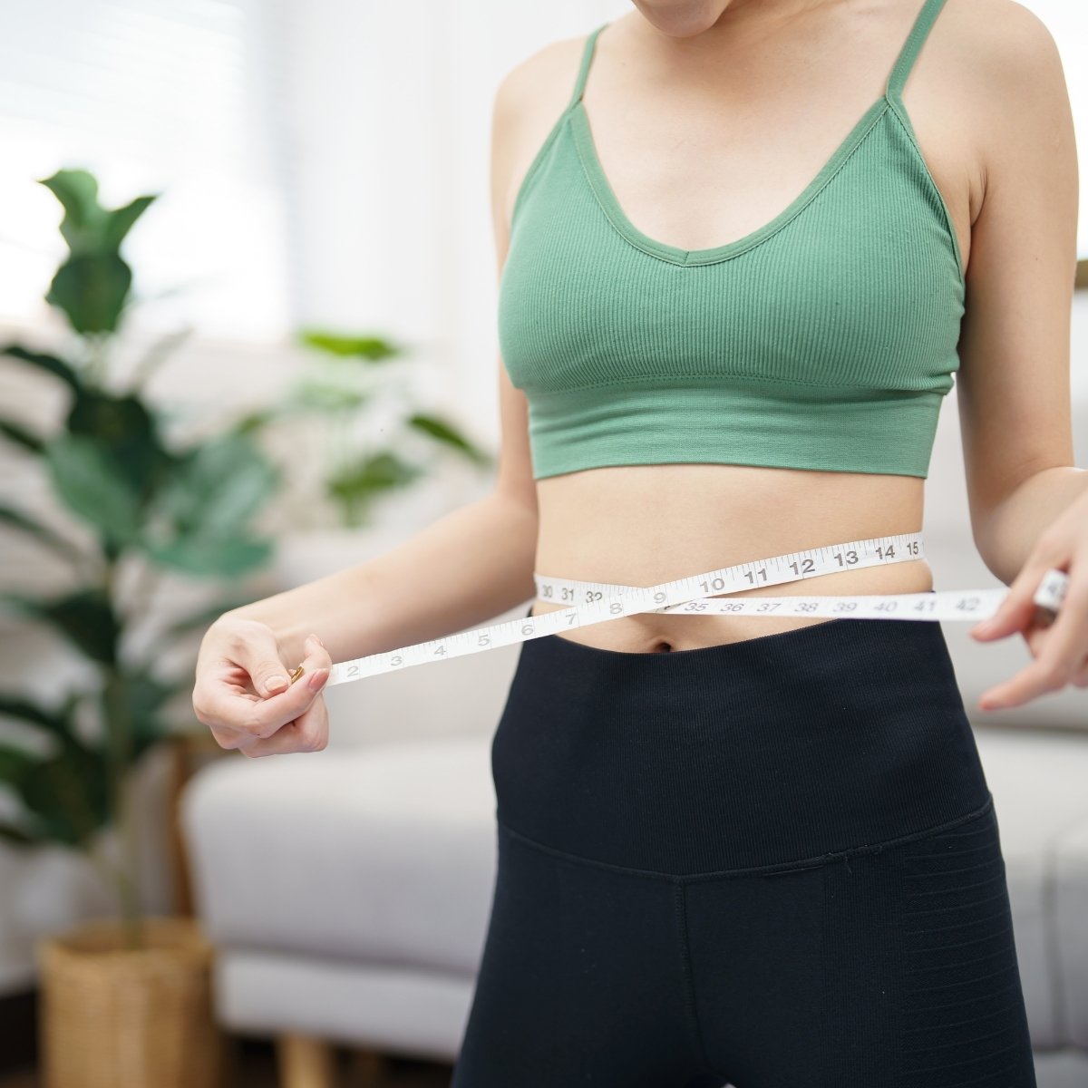 A person wearing a green sports bra and black leggings is holding a measuring tape around their waist, symbolizing weight management or tracking progress in a fitness journey.