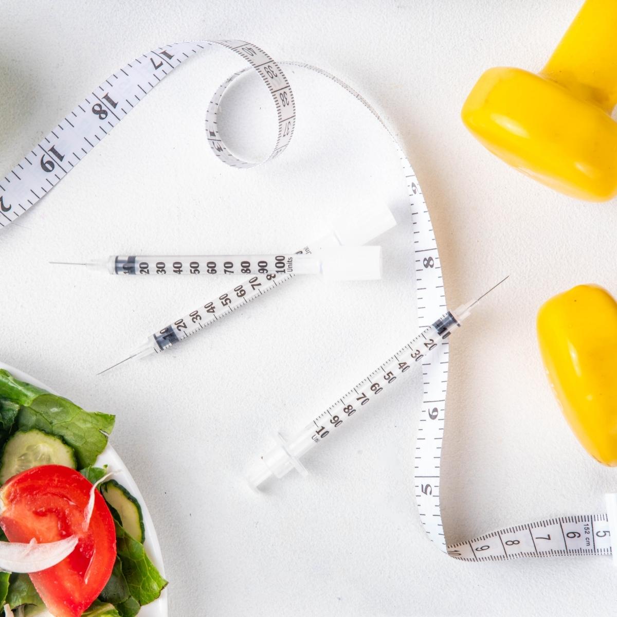 A flat lay image featuring three medical syringes, a measuring tape, a yellow dumbbell, and a salad bowl with fresh vegetables. The setup suggests themes of weight management, fitness, and health.