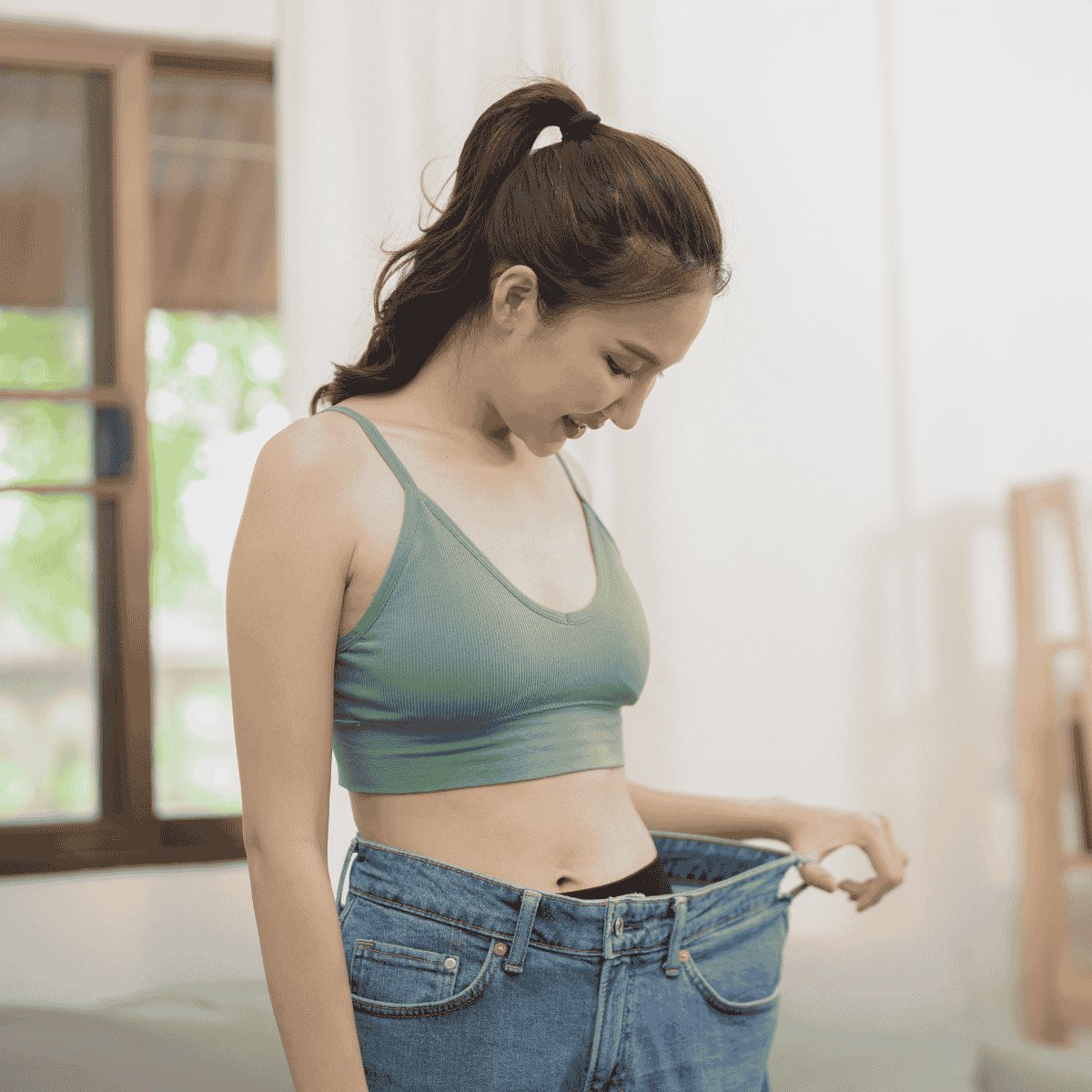 Woman in athletic wear smiling and holding out loose jeans after weight loss.