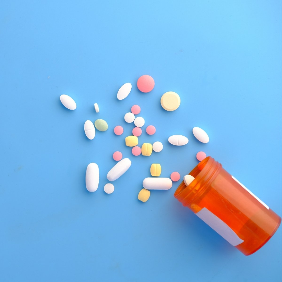 An assortment of colorful pills and tablets, including white, pink, yellow, and green, scattered on a light blue surface. An orange prescription bottle lies open with some pills spilling out.
