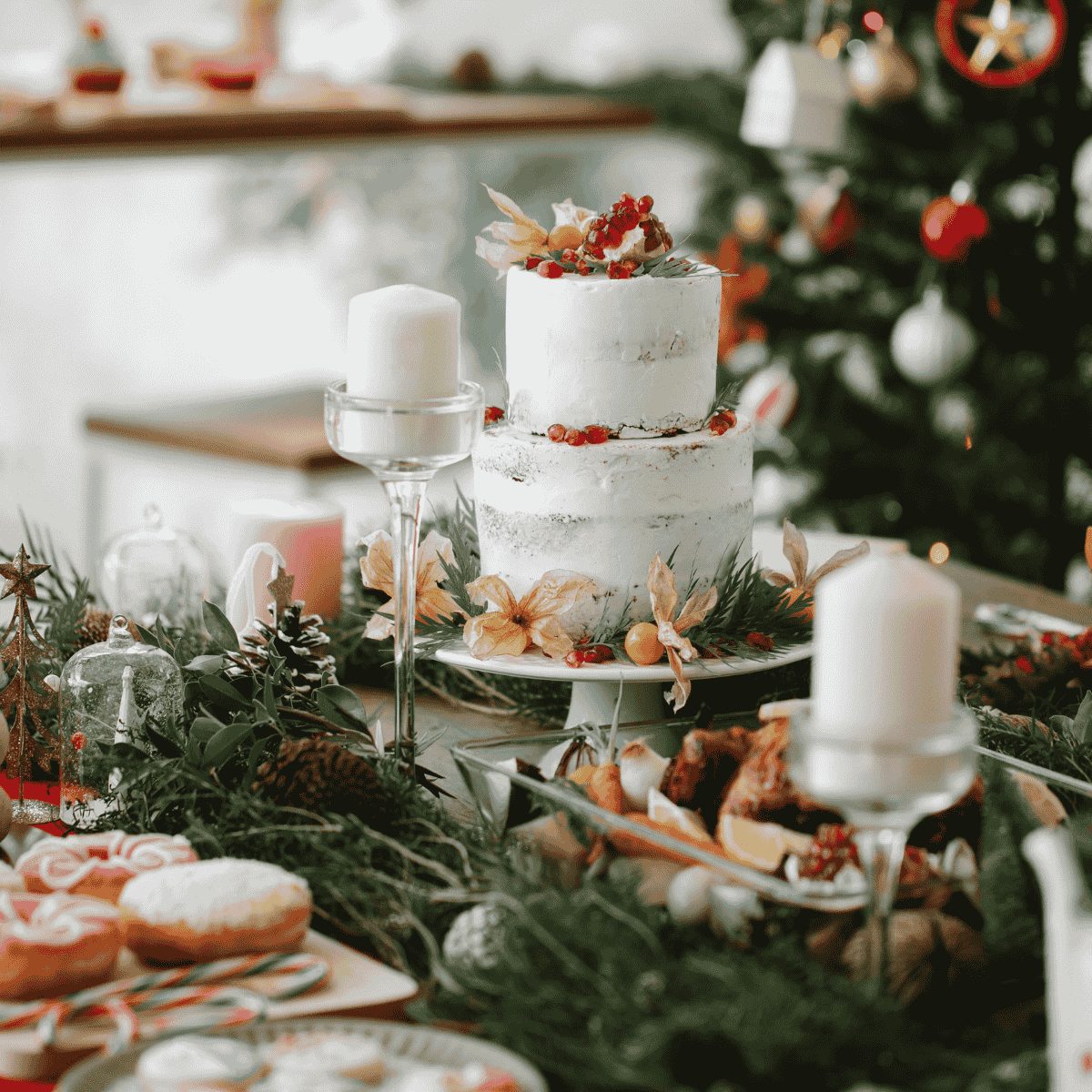 Festive holiday table setting with a two-tiered white cake decorated with red berries and greenery as the centerpiece. The table is adorned with candles, pine branches, pastries, and holiday treats, with a decorated Christmas tree in the background.